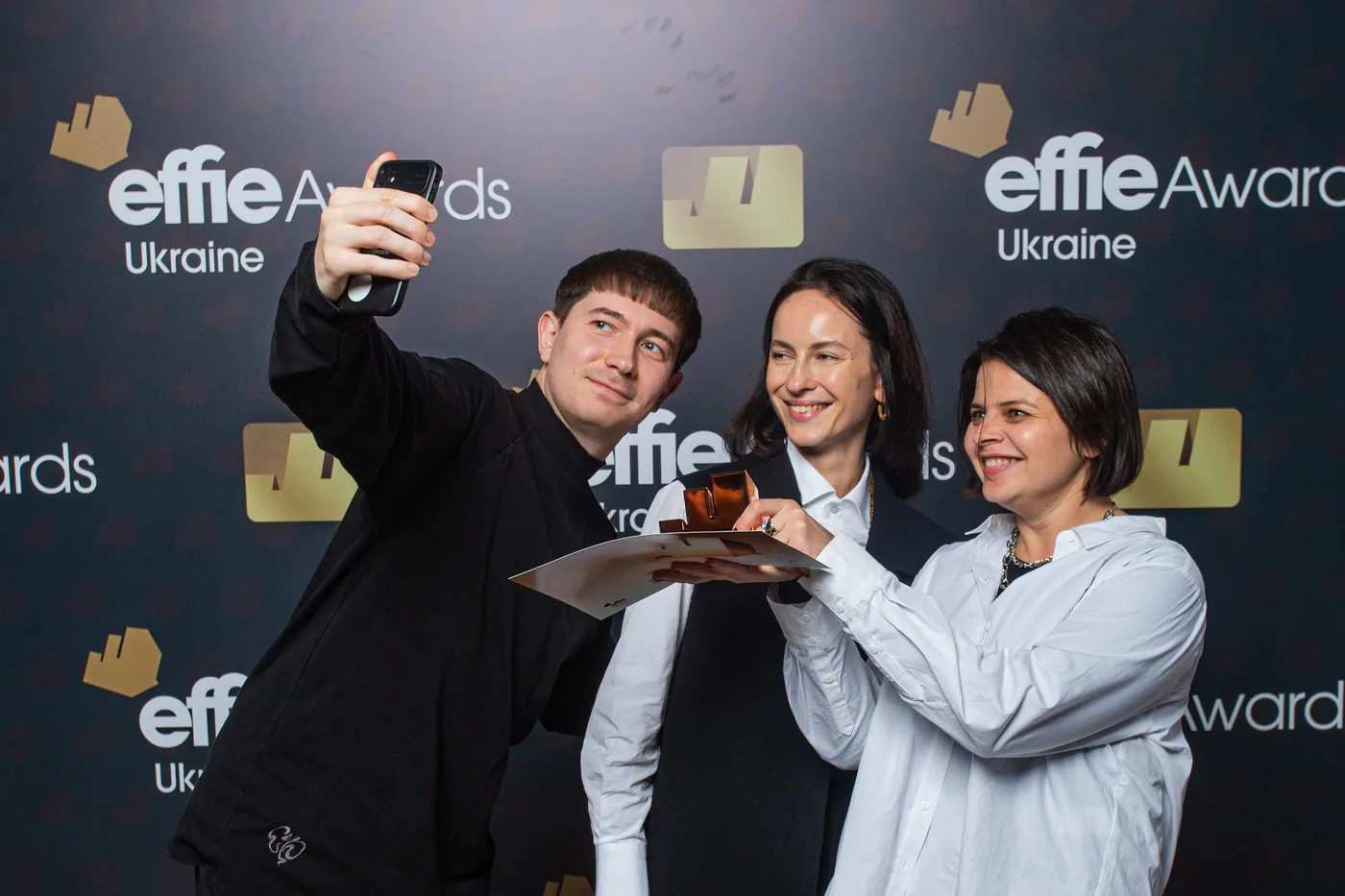 Three colleagues taking a selfie with a trophy in front of the Effie Awards Ukraine backdrop
