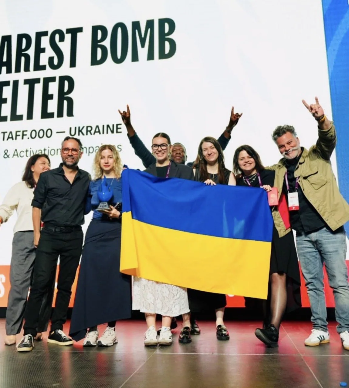 The team in front of the Nearest Bomb Shelter screen on stage, holding a Ukrainian flag